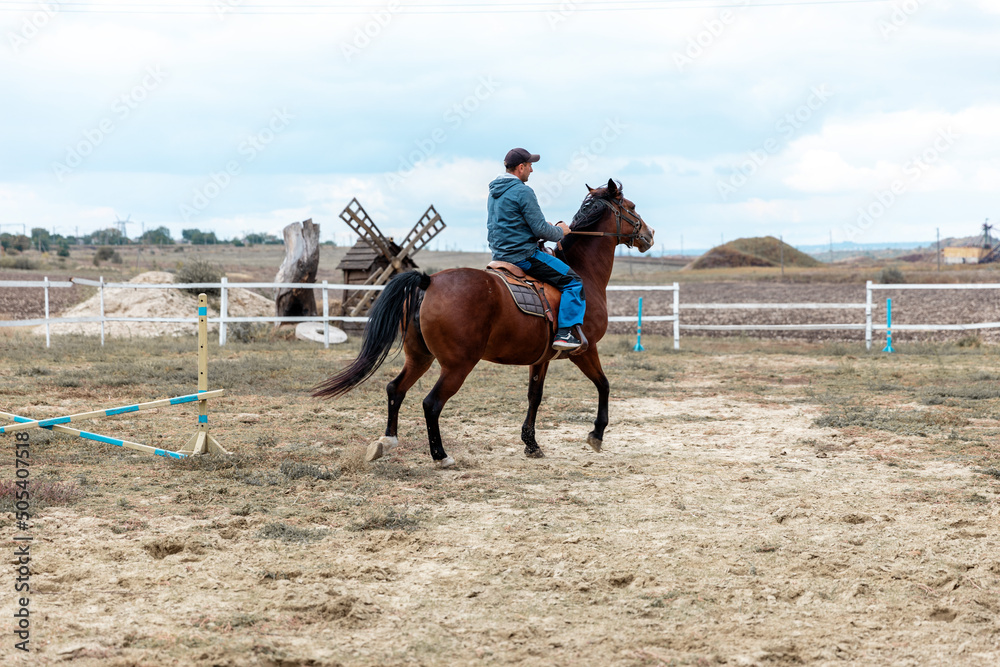 Φωτογραφία Back portrait of rider riding bay horse on village background with old ancient wooden mill