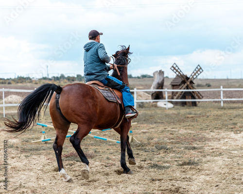 Rider riding bay horse on village background with old wooden mill. Back portrait