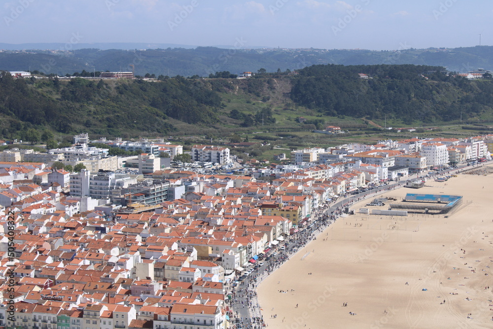 View over the city and the sandy beach of Nazaré. Lots of people walking by the beach. Mountain 
background and blue blured sky.