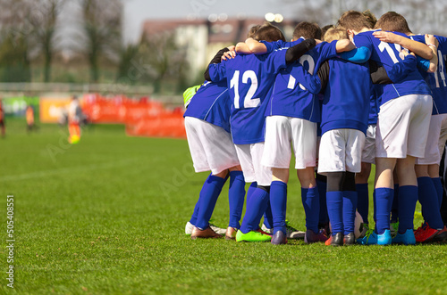 Kids soccer team with coach in group huddle before the match. Elementary age children are listening together to coach motivational speech. Boys in blue football shirts