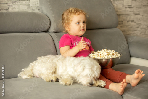 Cute little blond toddler girl, sitting in front of the TV with popcorn, watching movie with her pet dog