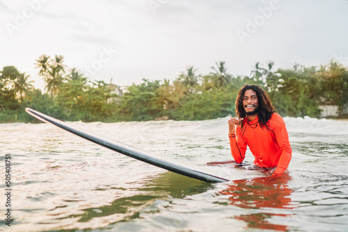 Fototapeta Naklejka Na Ścianę i Meble -  Black long-haired teen man showing shaka sign floating on long surfboard, waiting wave ready surfing with palms litted sunset rays. Extreme water sports or traveling to exotic countries concept
