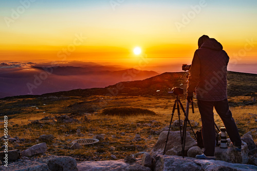 Man with camera on tripod take photo from sunset above clouds