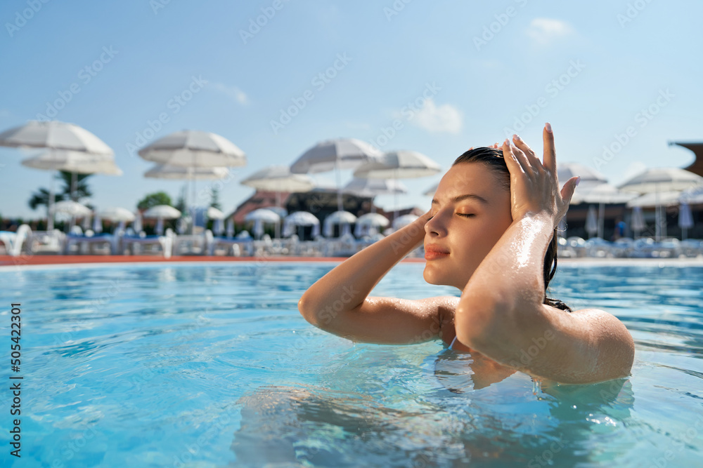 Side view of pretty young female swimming in pool. Slim brunette girl standing with closed eyes in water, touching hair, relaxing, enjoying. Concept of vacation and youth.