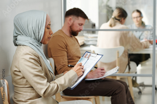 Young adult Caucasian man and Muslim woman in hijab sitting on chairs in visa service agency filling in application forms while waiting for appointment photo