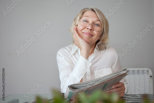 Smiling elegant woman in glasses sitting at a glass table looking at the camera and holding a newspaper in her hands. Satisfied with what I read.