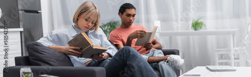 young blonde woman and african american man reading books near gadgets in modern living room, banner.