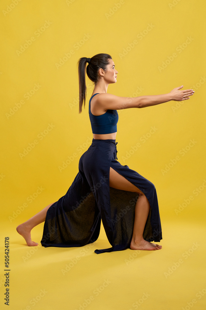 Yogi girl with ponytail keeping arms stretched, while staying in yoga pose indoor. Side view of fit woman in wide pants, practicing yoga, isolated on orange studio background. Concept of yoga. Yogi girl with ponytail keeping arms stretched, while staying in yoga pose indoor. Side view of fit woman in wide pants, practicing yoga, isolated on orange studio background. Concept of yoga.