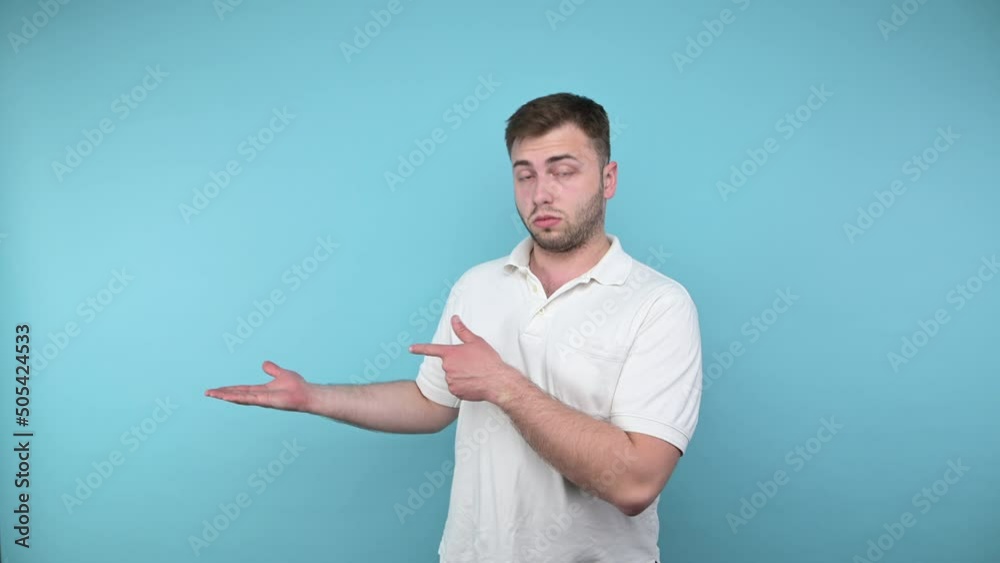 Portrait of a surprised man in a white T-shirt on a blue background looks at the camera with a shocked face and points his hand to the side on copy space