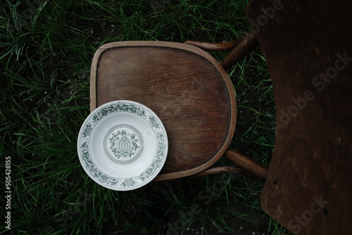 Grandma's white kitchen bowl with a green floral pattern on an old wooden chair in the garden. Cottage core concept. Dark photo. Top view. Copy space.   © Ganna Zelinska