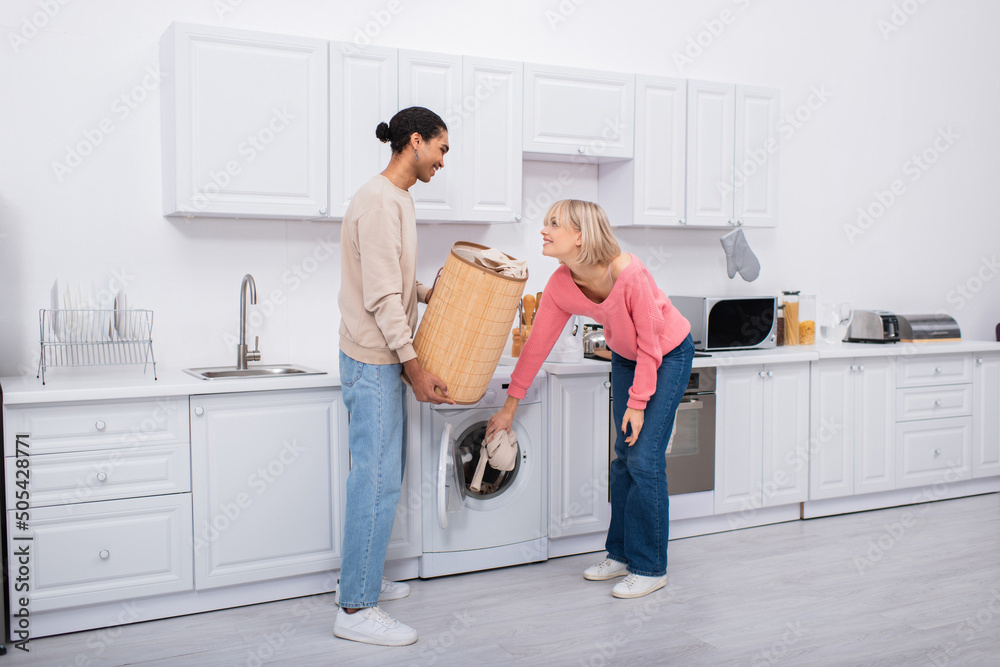 Φωτογραφία happy blonde woman putting clothes in washing machine near african american man