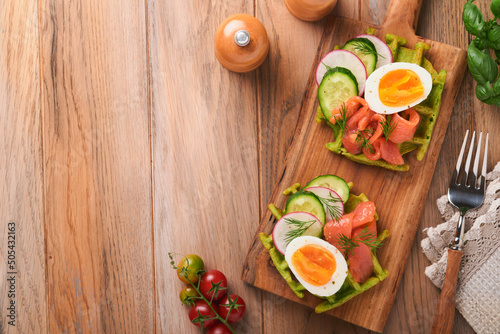 Savory waffles. Breakfast green waffles wild garlic or spinach with egg  tomato  salmon on wooden plate for breakfast on old wooden background. Light dinner  lunch or breakfast. Top view. Mock up.