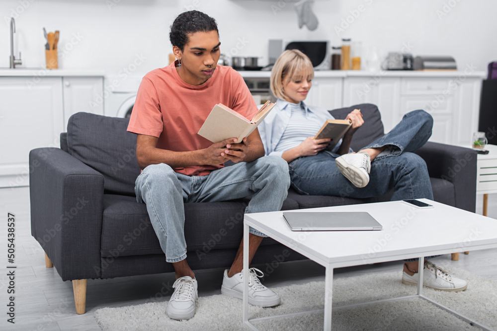 young african american man and blurred woman reading books near gadgets on coffee table.