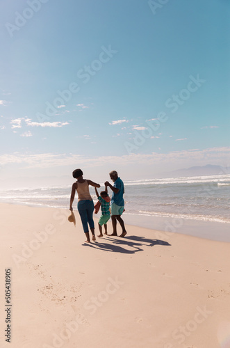 African american grandparents holding grandson's hands and walking at sandy beach against blue sky