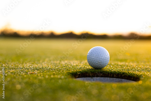 Close-up of white golf ball by hole on grassy land against clear sky during sunset, copy space