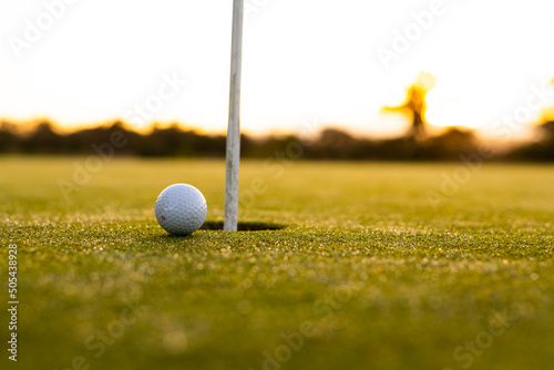 White golf ball by hole and flagstick on grassy land against clear sky during sunset, copy space