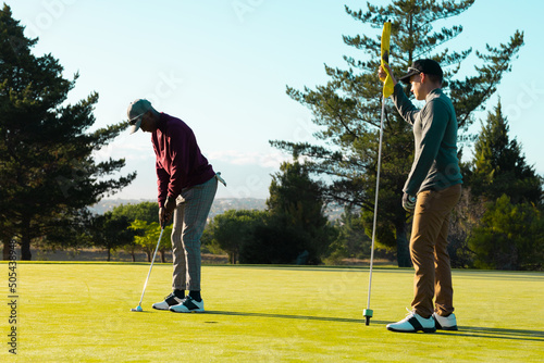 Caucasian young man holding golf flag while african american friend hitting golf ball with club