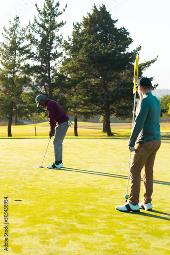 Caucasian young man with golf flag and african american friend hitting golf ball with club at course