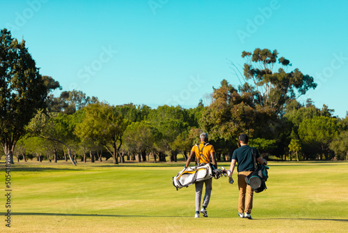 Rear view of multiracial young male friends with golf bags walking at golf course against clear sky