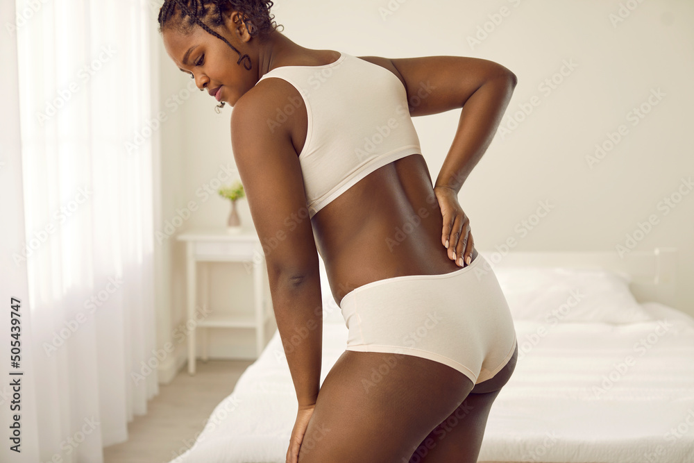 Foto de Black woman suffering from backache or kidney pain. Plus size young  African lady in white underwear standing in bedroom at home, leaning  forward and holding hand on lower back. Health