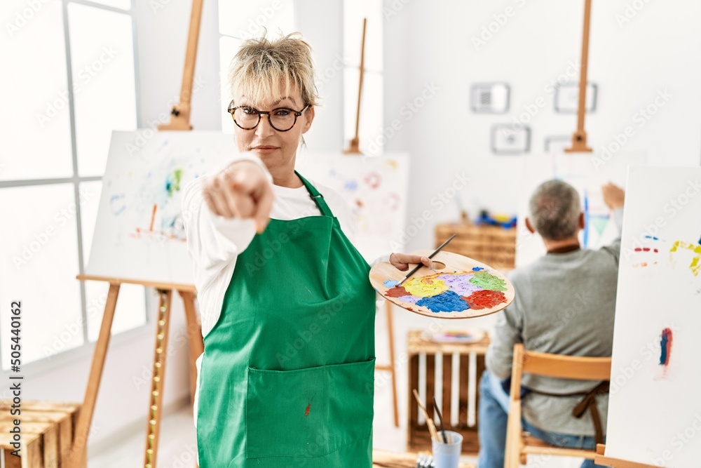 Middle age caucasian woman drawing canvas at art studio pointing with finger to the camera and to you, confident gesture looking serious Middle age caucasian woman drawing canvas at art studio pointing with finger to the camera and to you, confident gesture looking serious