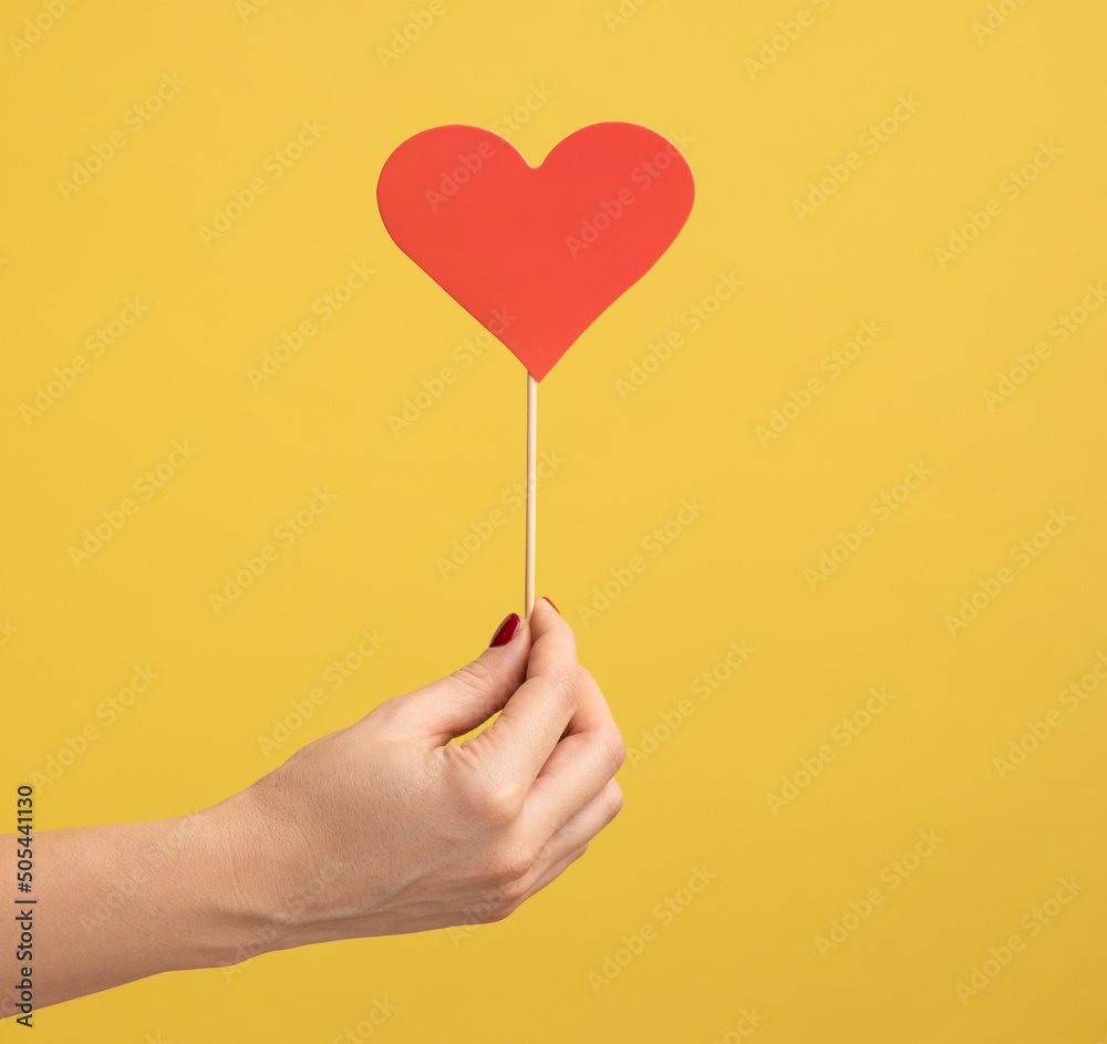 Profile side view closeup of woman hand holding small red heart stick in hand, symbol of love and gentle. Indoor studio shot isolated on yellow background. Profile side view closeup of woman hand holding small red heart stick in hand, symbol of love and gentle. Indoor studio shot isolated on yellow background.