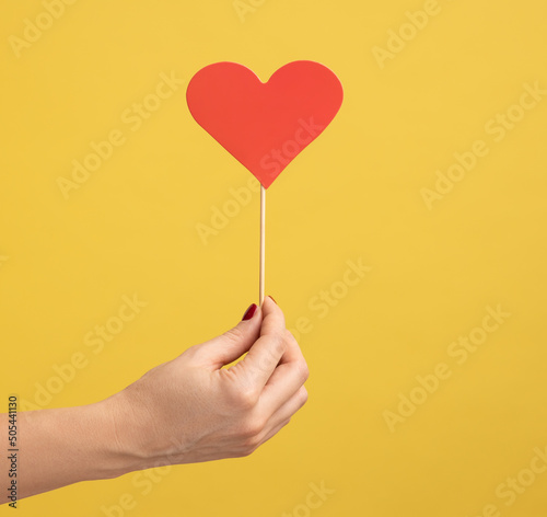 Profile side view closeup of woman hand holding small red heart stick in hand, symbol of love and gentle. Indoor studio shot isolated on yellow background.
