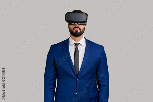 Bearded man standing in vr headset, watching virtual reality movie with calm facial expression, innovative technology, wearing official style suit. Indoor studio shot isolated on gray background.