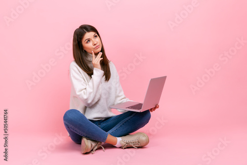 Pensive female sitting on floor with crossed legs, holding notebook, thinking about new project, wearing white casual style sweater. Indoor studio shot isolated on pink background.