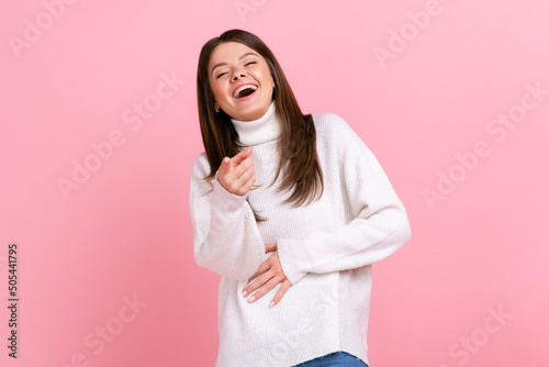 Photos Happy female laughing out loud and pointing finger to camera, hearing funny joke, holding belly, wearing white casual style sweater