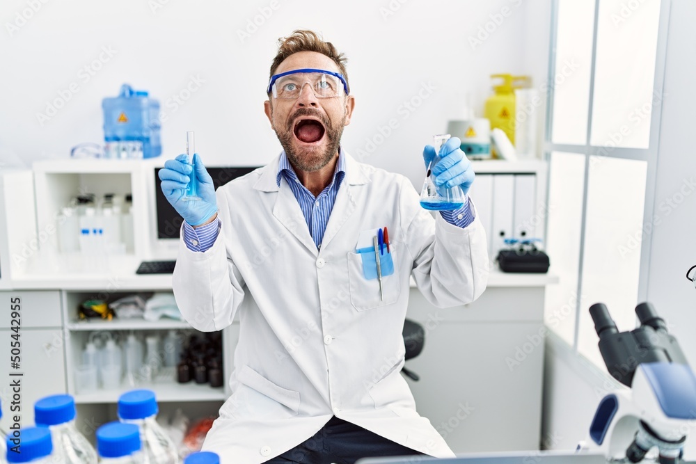 Middle age man working at scientist laboratory holding chemical products angry and mad screaming frustrated and furious, shouting with anger looking up.