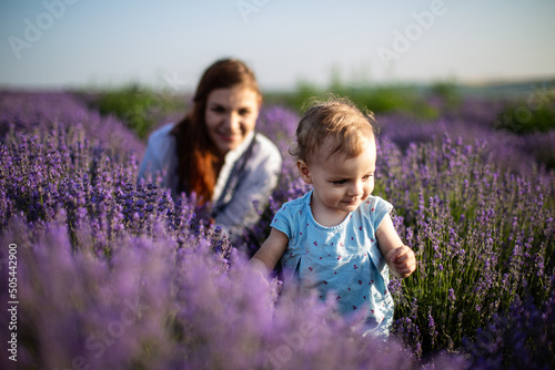 Cute smiling baby girl with blonde curly hair wearing a blue dress walking on a lavender field. Mother and baby daughter having fun on the lavender field