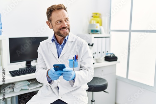Middle age hispanic man wearing scientist uniform using smartphone at laboratory