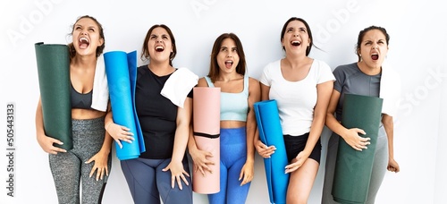 Group of women holding yoga mat standing over isolated background angry and mad screaming frustrated and furious, shouting with anger. rage and aggressive concept.