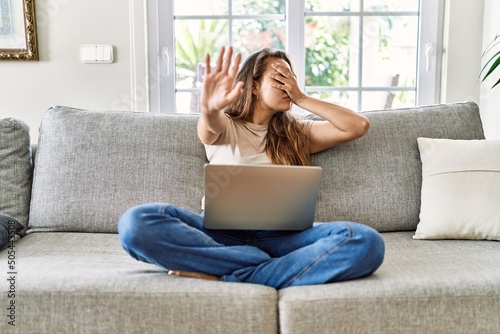 Fotografering Beautiful young brunette woman sitting on the sofa using computer laptop at home covering eyes with hands and doing stop gesture with sad and fear expression