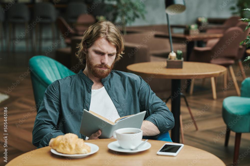 Serious handsome young man with beard sitting at table and calling colleague by phone while waiting for him in cafe Serious handsome young man with beard sitting at table and calling colleague by phone while waiting for him in cafe