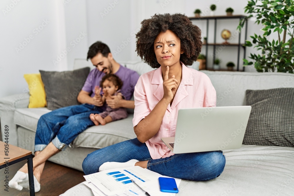 Mother of interracial family working using computer laptop at home thinking concentrated about doubt with finger on chin and looking up wondering Mother of interracial family working using computer laptop at home thinking concentrated about doubt with finger on chin and looking up wondering