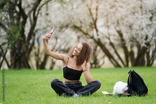 Cute african american teen girl using smartphone and enjoying the blooming spring nature in a sunny day      