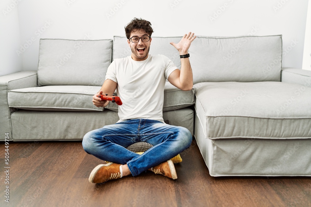 Hispanic man playing video game holding controller by the sofa celebrating victory with happy smile and winner expression with raised hands Hispanic man playing video game holding controller by the sofa celebrating victory with happy smile and winner expression with raised hands