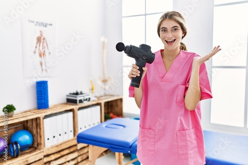 Young physiotherapist woman holding therapy massage gun at wellness center celebrating victory with happy smile and winner expression with raised hands