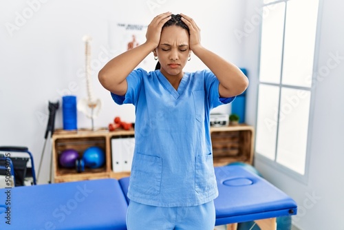 Young african american woman working at pain recovery clinic suffering from headache desperate and stressed because pain and migraine. hands on head.