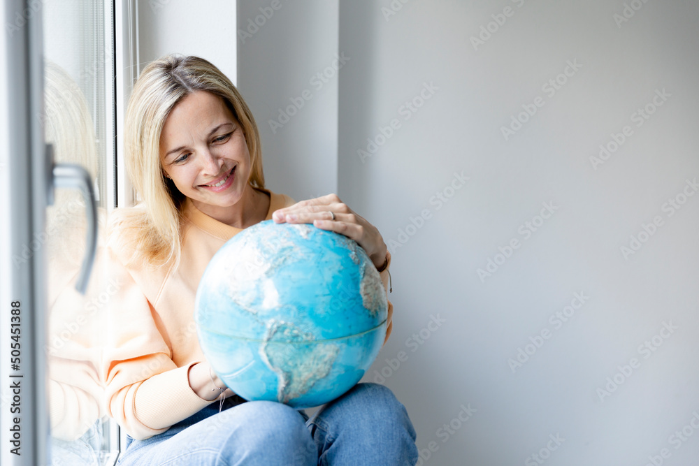 young blonde woman sitting at window sill holding globe and dreaming about vacation, freedom, leave, travelling young blonde woman sitting at window sill holding globe and dreaming about vacation, freedom, leave, travelling