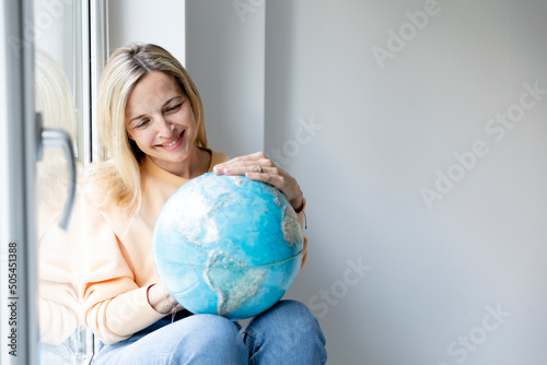 young blonde woman sitting at window sill holding globe and dreaming about vacation, freedom, leave, travelling