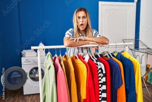 Young blonde woman at laundry room with clean clothes in shock face, looking skeptical and sarcastic, surprised with open mouth