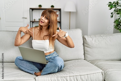 Hispanic woman sitting on the sofa at home using laptop looking confident with smile on face, pointing oneself with fingers proud and happy.