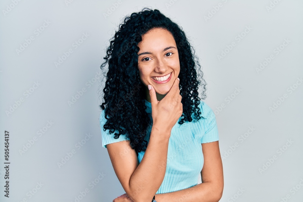 Young hispanic woman with curly hair wearing casual blue t shirt looking confident at the camera smiling with crossed arms and hand raised on chin. thinking positive.