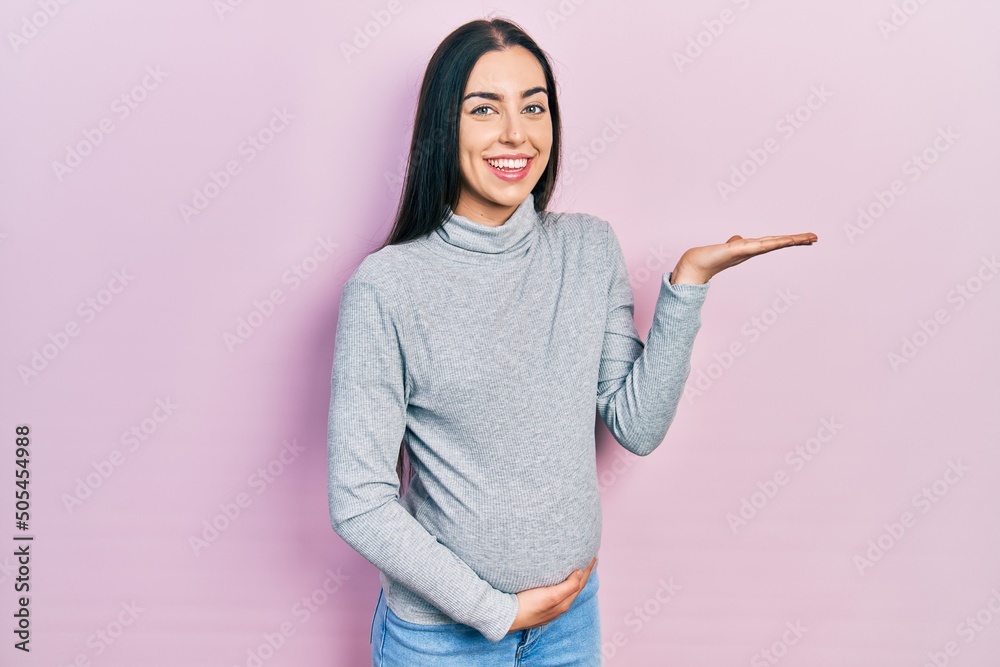 Beautiful woman with blue eyes expecting a baby, touching pregnant belly smiling cheerful presenting and pointing with palm of hand looking at the camera. Beautiful woman with blue eyes expecting a baby, touching pregnant belly smiling cheerful presenting and pointing with palm of hand looking at the camera.