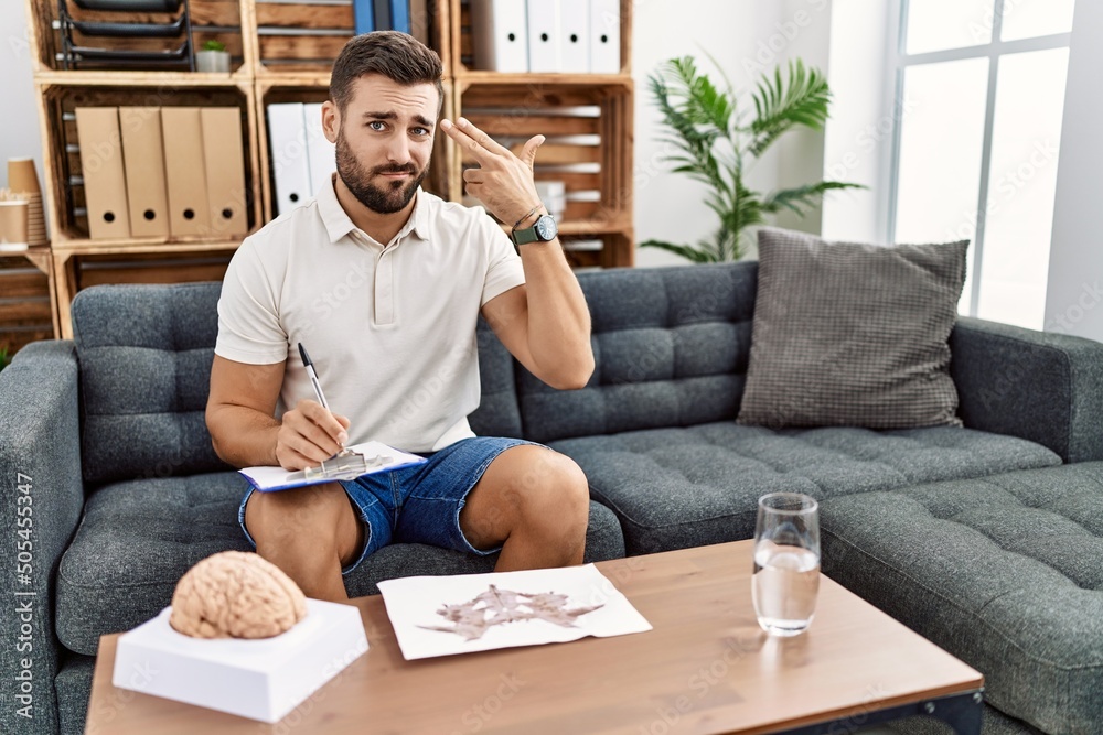 Handsome hispanic man working with rorschach test at psychology clinic shooting and killing oneself pointing hand and fingers to head like gun, suicide gesture. Handsome hispanic man working with rorschach test at psychology clinic shooting and killing oneself pointing hand and fingers to head like gun, suicide gesture.