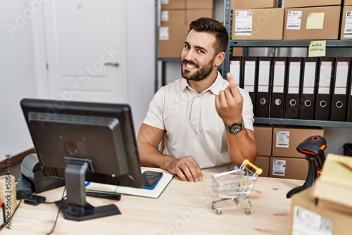 Handsome hispanic man working at small business commerce beckoning come here gesture with hand inviting welcoming happy and smiling