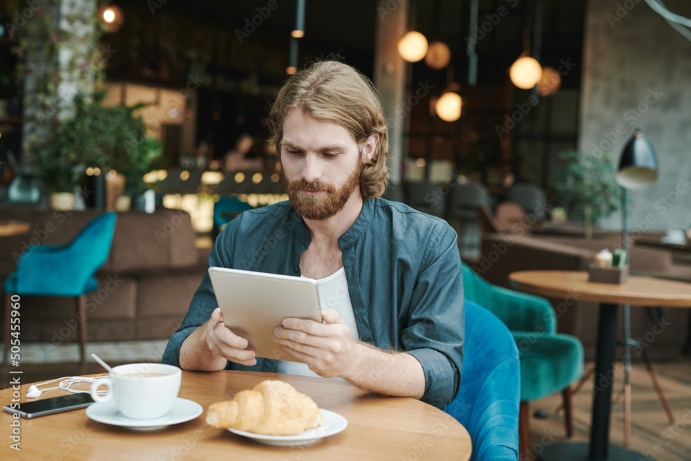 Concentrated young bearded man in casual shirt drinking coffee and eating croissant while checking smartphone in cafe